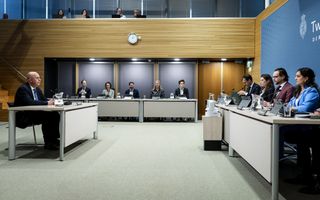 Man zit achter tafel om verhoord te worden door enquêtecommissie. Ook de commissieleden zijn in beeld: vier zitten er voor hem, vijf zitten er links naast hem.