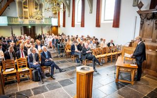 Een man in zwart pak, bril, spreekt de aanwezigen toe. Op de voorgrond het doopvont. Achterin de kerk het orgel.