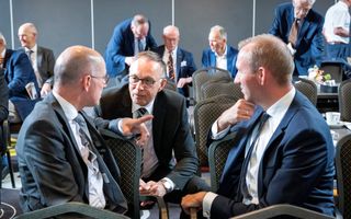 Drie mannen zijn zittend met elkaar in gesprek. Op de achtergrond staan en zitten andere mensen.