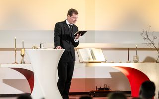 Een man in zwart pak leest uit een Bijbel voor bij een statafeltje.