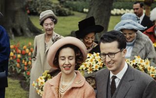 Een man in een grijs pak houdt de arm van een vrouw in oranje mantelpakje vast. Zij draagt narcissen.