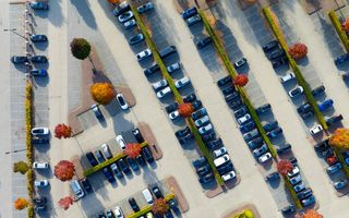 Een parkeerplaats van boven gezien. Strakke groene heggen scheiden de vakken in de lengte, rode boompjes torenen daarboven uit. Overheersende autokleur is zwart, gevolgd door allerlei tinten grijs.
