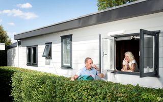 Achter de heg drinkt een man in blauwe polo staand een kop koffie. Een vrouw leunt naast hem uit het raam van een stacaravan.