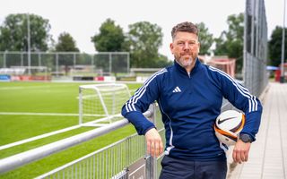 Man met blauw trainingsjack staat in ontspannen houding naast een voetbalveld. 