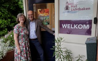 Een vrouw, een bril op, grijs haar en een bloemetjesjurk aan, en een man, een bril op, kaal en een grijs colbert aan bij de deur, met daarnaast de woorden: Het Lichtbaken. Welkom.