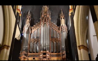 Het front van een oud orgel hangt in een kerk met overwegend donkere wanden. Bovenop staan houten beelden.