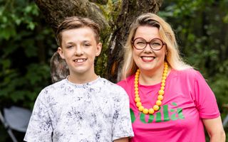 Een tienerjongen en een volwassen vrouw poseren voor een boom in een groene omgeving. De vrouw draagt een felroze shirt en een oranjegele kralenketting.