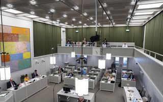 Een vergaderzaal met meerdere rijen afzonderlijke bureaus waar mensen zitten te werken of vergaderen. Bovenin is een publieke tribune met enkele toeschouwers, en links aan de muur hangt een groot kleurig schilderij.