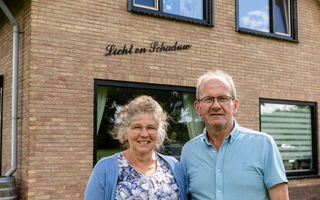 Echtpaar, beiden in het blauw en met een bril, voor hun huis waarop in losse metalen letters de naam Licht en Schaduw staat.