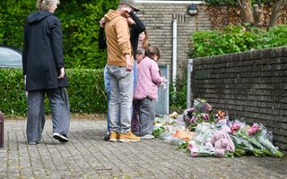 Enkele personen staan op een stoep naast een lage muur en kijken naar een plek waar veel bloemen, boeketten en knuffels op de grond liggen. Op de achtergrond staan huizen en groen.