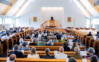 Ds. Fokkema, een man in zwart kostuum, spreekt in een kerk; mensen zitten in houten kerkbanken. 