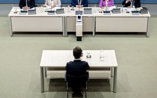 Drie mannen en twee vrouwen zitten achter een lange tafel. Voor hen een man in een donker pak eenzaam achter een losse tafel. 