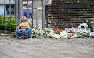 Een klein meisje in een bruin jasje, met een spijkerbroek en het haar in een paardenstaart, legt bloemen bij een ijzeren hek. Rechts van het hek een muur waarvoor ook veel bloemen liggen. Op de achtergrond, door het hek te zien, een schoolplein.