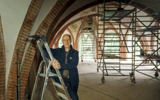 Een oudere man, op een trapje in een kerk. De kerk staat vol steigers.