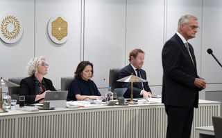 Twee vrouwen en een man in pak zitten achter een wit bureau. Voor hen staat een man in een donker pak.