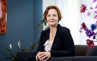 Glimlachende vrouw in donkerblauw, zittend in fauteuil met handen op elkaar.