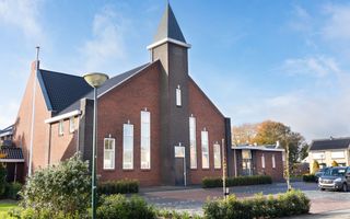 Een nieuwe kerk, met een torentje, ervoor parkeerplaatsen. De lucht is blauw.