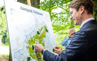 Een man met bril in blauw kostuum staat voor een bord met een grote kaart van de Veluwe. Op het bord staat: Aanpak Veluwe.