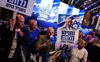 Vier mannen, van wie een met een luidspreker, dragen een spandoek met het gezicht van premier Netanyahu erop. Twee dragen borden met Hebreeuwse tekst. 