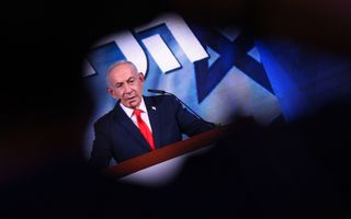 Man in donker pak en met rode stropdas staat achter spreekgestoelte.