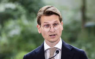 Een man in een pak staat achter een microfoon, voor een groene achtergrond.