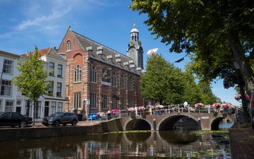 Fraai oud gebouw met torentje aan gracht met bomen en een stenen boogbrug.