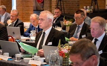 Mannen in donkere pakken, achter vergadertafels. Voor hen koffiekopjes, waterkannen, laptops en bloemstukjes.