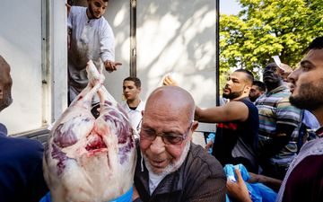 Een oudere man met een kort baardje heeft een groot stuk vlees, dat half uit een blauwe plastic zak steekt, in zijn armen. Op de achtergrond andere jonge mannen.
