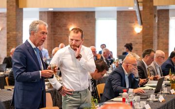 Mannen achter lange vergadertafels, lezend in papieren en kijkend naar hun beeldscherm. Op de voorgrond twee mannen die staan en die met elkaar in gesprek zijn. 