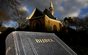 Op de voorgrond een Bijbel, op de achtergrond een verlicht kerkje tegen de avondhemel.