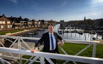 Eddy Bilder, met donker jasje en stropdas. Op de achtergrond bootjes in een haven.