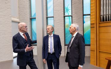 Drie heren in donkere pakken staan bij elkaar en voeren een gesprek. Rechts van hen staat een groot pijporgel.