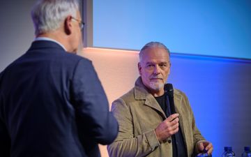 Man in bruinachtige jas, met microfoon in de hand.