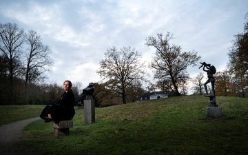 Vrouw in zwarte jas zit op een stenen sokkel op een grasveld. Ernaast twee sokkels met zwarte standbeelden.