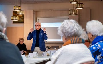 Oudere vrouwen luisteren naar een man in blauw geruite colbert.