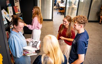 Een vrouw met een spijkerjurkje aan en een bril op houdt een stapel boeken vast. Rechts van haar een groepje jongeren dat naar de boeken kijkt.