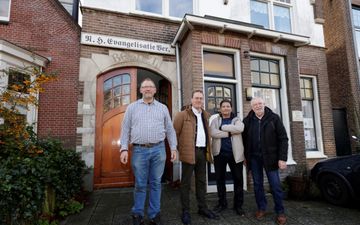 Vier mannen staan op de stoep van een gebouw uit de jaren ‘20 van de vorige eeuw. Het gebouw heeft houten deuren in de stijl van art deco en een erkertje. 