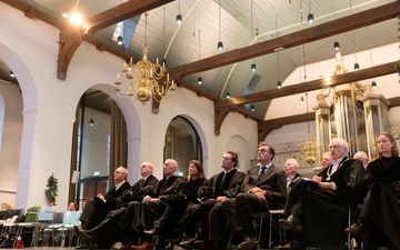 Hoogleraren in zwarte toga kijken naar omhoog naar de man op het podium. Achter hen het orgel.