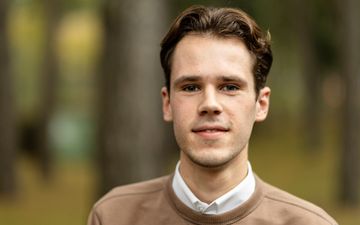 Een jongeman met bruin haar, gekleed in een wit overhemd met daarover een bruine trui.