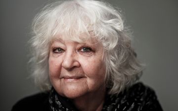 Foto van een oude vrouw met korte, grijze krullen. Ze draagt een zwart vest met witte stiksels. De achtergrond is grijs. 