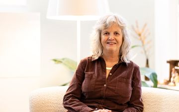 Een vrouw met grijs haar zit voor een lamp op een witte stoel.