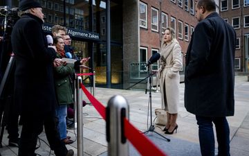 Blonde jonge vrouw in witte winterjas praat op een plein tegen journalisten, die achter een rood lint staan.