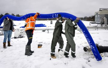 Vier mannen in de sneeuw dragen met zijn allen een blauwe slang. Ze staan op een dak en ontdoen het van sneeuw. 
