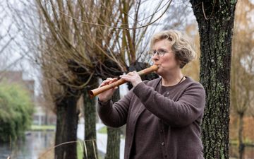 Een vrouw van middelbare leeftijd speelt buiten op een blokfluit. Achter haar zijn knotwilgen en een sloot te zien.