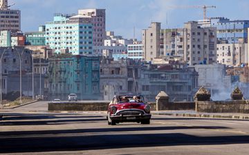 Een rode auto rijdt over een bijna lege asfaltweg. Op de achtergrond is hoogbouw te zien.