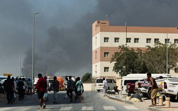 Mensen lopen op straat, terwijl op de achtergrond donkergrijze rookwolken opstijgen.