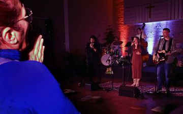 Een vrouw in een blauw shirt heeft haar handen gevouwen. Op de achtergrond is een muziekgroep met twee zangers, een gitarist, contrabassist en drummer te zien.