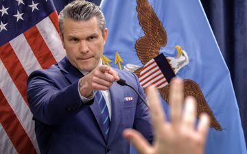 Man met grijs haar en blauw pak voor de Amerikaanse vlag die wijst naar een opgestoken hand