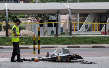 Een man met een petje op en een hesje aan staat naast de brokstukken van een grijze drone.