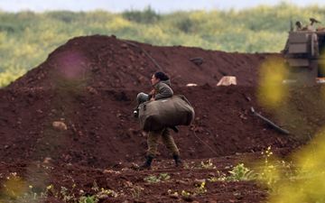 Vrouw in legeruniform draagt bepakking langs een aarden wal.